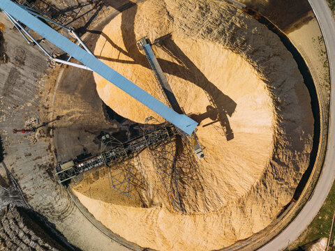 Aerial view of a vast, golden wood chip mountain, starkly textured, with a prominent blue conveyor casting a deep shadow, Cacia, Aveiro District, Portugal.