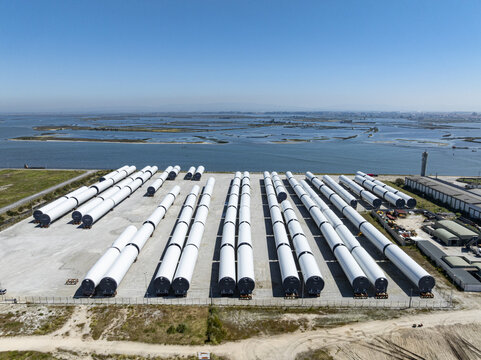 Aerial view of neatly arranged wind turbine blades casting shadows on the industrial ground contrasting with the distant water, Aveiro, Aveiro, Portugal.