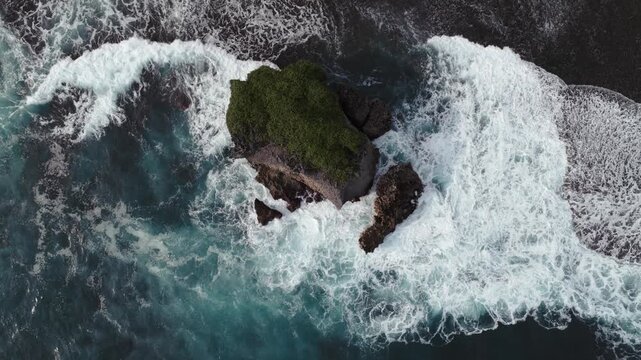 Aerial view of turquoise ocean waves crashing against the rocky cliffs of Mesra Beach with white sea foam and green vegetation Kecamatan Tanjungsari, Daerah Istimewa Yogya, Indonesia.