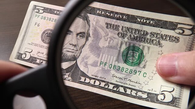 Examination of 5 American dollar (USD) cash money with magnfication glass, loupe. Concept of authenticity check, fight against counterfeiting of banknotes, anti fraud measures in USA. Close up view.