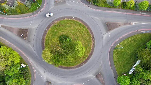 Aerial top-down view of vehicles driving around a roundabout in the United Kingdom with left-hand traffic. Cars moving smoothly through circular road junction surrounded by greenery.