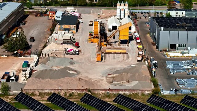 Aerial view of an industrial yard with aggregate piles, concrete production equipment, warehouse buildings, and solar panels along the edge of the site.