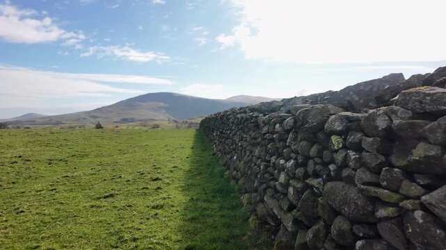 Dry stone wall separating lush green grass under a blue sky in English Cumbrian landscape. POV, push forward shot