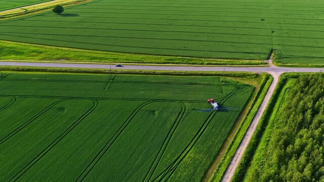 Red tractor with wide boom crop sprayer moves across vast green grain fields leaving geometric track patterns in aerial drone footage over rural Latvia