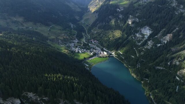 Aerial View Of Lago di Isola In Madesimo, Italy, Surrounded By Pine-forested Slopes And Village. pullback shot