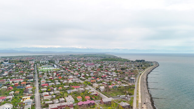 Aerial view of potis black sea shoreline with breakwater, green promenade and mixed rooftops, urban coastline framed by cloudy skies and distant snow capped mountains