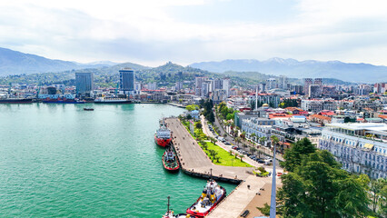 Obraz premium Batumi cityscape with bustling black sea harbor, mix of modern and historic waterfront architecture, ships and boats, and snow capped caucasus mountains on clear day