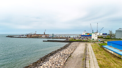 Obraz premium Industrial port showing a cargo ship docked, cranes loading containers, breakwaters, and various port infrastructure, representing international trade and the global shipping industry