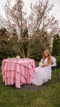 Young woman in white dress reads book at pink tea party table under blooming magnolia tree