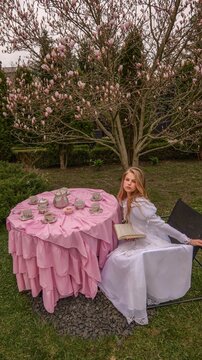 Young girl in white dress reads book at pink tea party table under blooming magnolia tree