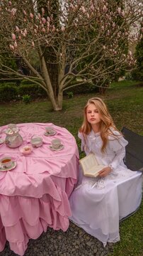 Young girl in white dress reads book at a pink tea party table outdoors
