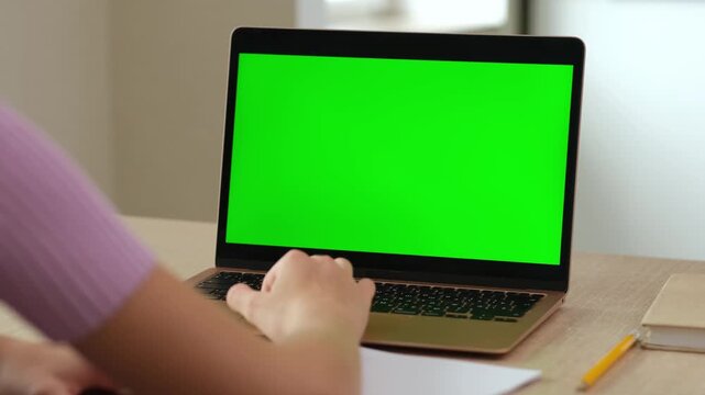 Back view of teen girl typing on keyboard of laptop with green screen. Female person using chroma key computer blank mock up at home