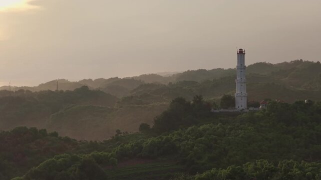 Aerial view of the tall white lighthouse standing on a verdant hill overlooking the misty coastal landscape near Mesra Beach Kecamatan Tanjungsari, Daerah Istimewa Yogya, Indonesia.