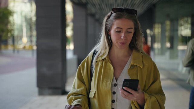 Young blonde woman traveler rushing through airport with suitcase using mobile phone to find directions while running late