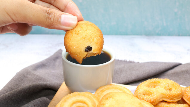 Close up of a hand dipping round butter cookies into a cup of black coffee, capturing a warm and cozy snack moment.