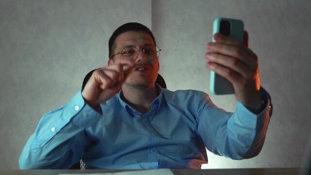 Man in blue shirt working late at desk in office, talking to client through video call on mobile phone. Concept of business meeting, planning, communication and productivity.