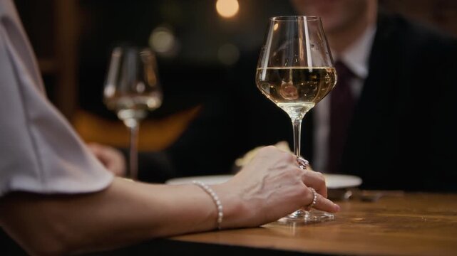 Cropped close up shot of hand of unrecognizable elegant woman wearing pearl bracelet holding glass of white wine enjoying romantic dinner in luxury restaurant