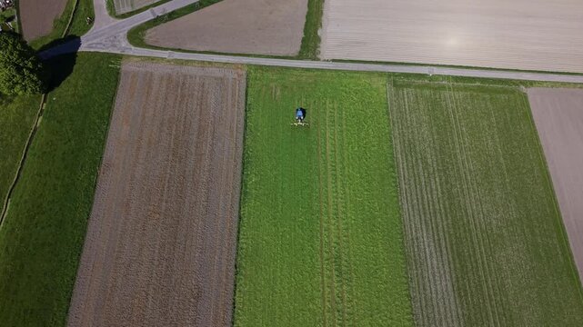 Aerial drone footage of a tractor turning cut grass in a farmland field, changing direction and moving toward the camera, showing active agricultural machinery at work during spring season.