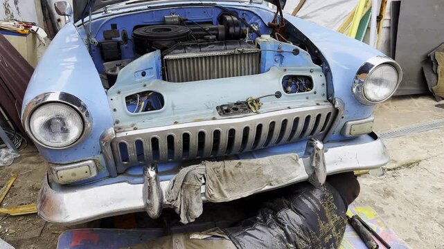 Male repairman lies under his vintage car at courtyard of house. Auto mechanic repairing his blue retro vehicle outdoor. Concept of automobile maintenance. Slow motion