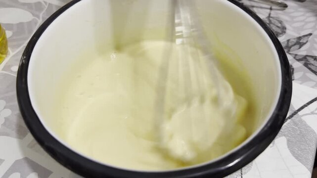 Female hand whisks batter in bowl on kitchen table preparing pancakes or fritters. The mixture becomes smooth and airy as beaten, with focus on the motion, texture and the homemade cooking process