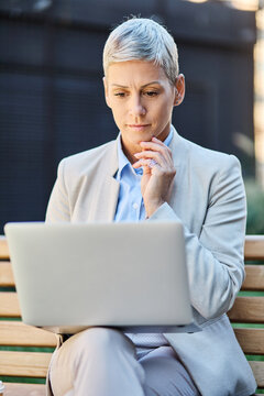 Portrait of a young businesswoman woman using a laptop computer and sitting on a bench in a coty park, surrounded by modern corporate office buildings