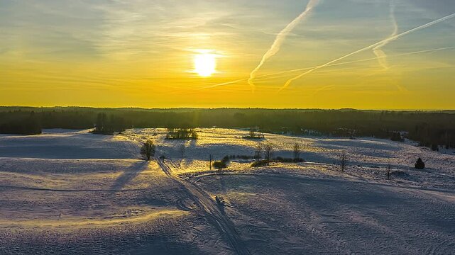 Winter landscape at sunset, golden hour light illuminating snow fields and bare trees in aerial timelapse