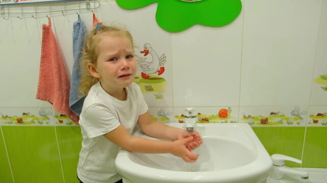 Moskow, Russia, 21.08.2025 Crying little girl washing hand in bathroom sink. Sad child standing at faucet with water. Emotional toddler hygiene routine in nursery school or home restroom.