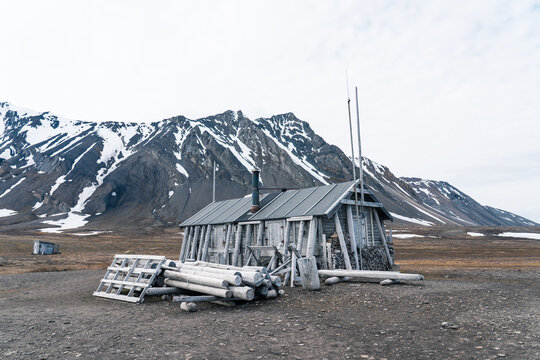 View of the historic Bamsebu cabin with piled logs against snow-capped mountains in the arctic tundra Svalbard, Svalbard and Jan Mayen.