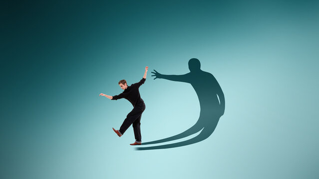 Man trying to keep balance while large shadow reaches toward him. Concept of inner fear and anxiety, psychological pressure and struggle with self perception and hidden emotions.