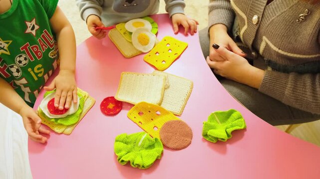 Moskow, Russia, 21.08.2025 Kid and woman playing with handmade crocheted toy food on pink table. Toddler learning to prepare sandwich using knitted ingredients. Educational game concept.