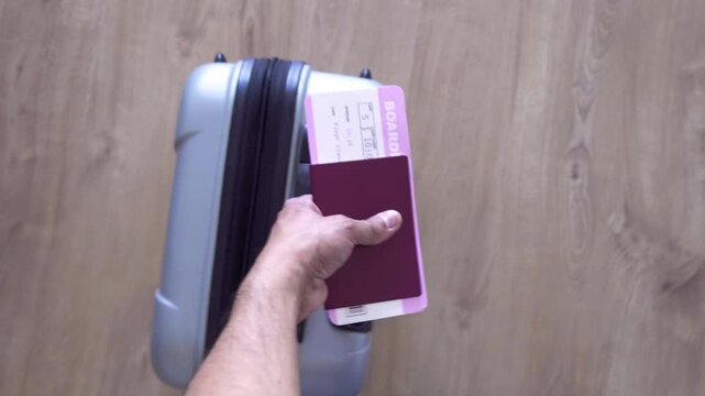 First-person perspective of a hand holding an passport and boarding pass, pulling a gray suitcase through an airport terminal.