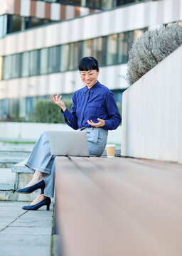 Portrait of a young asian japanese businesswoman woman using a laptop computer having a video call conference and sitting on a bench in a coty park, surrounded by modern corporate office buildings