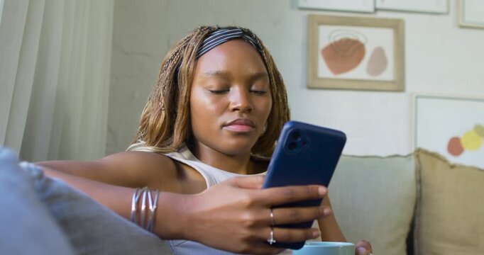 African American woman scrolling phone on couch with mug, noticing off-screen movement and reacting