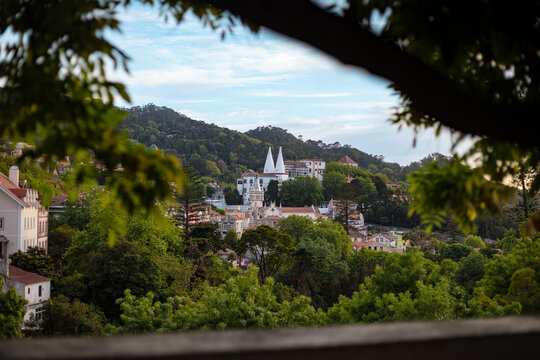 Sintra, Portugal - Framed view of the historic town of Sintra surrounded by lush green forest, featuring the iconic twin chimneys of the National Palace under a soft evening sky.