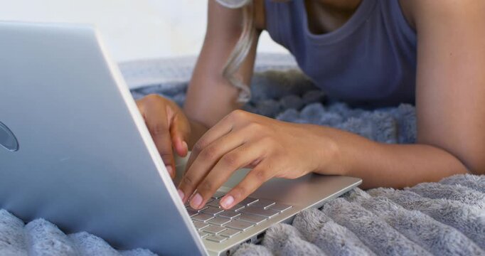 Adult African woman typing on silver laptop on bed, leaning forward, wearing headphones studying