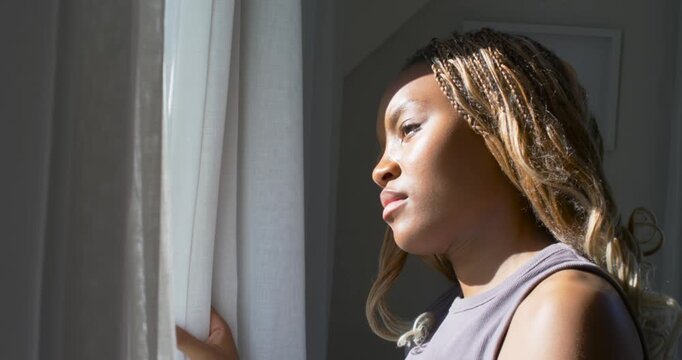 Noticing sun African-American woman holding curtain tilting head and gazing out window with braids