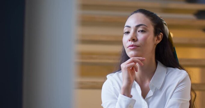 Woman in stairwell resting chin on right hand in blouse, stairs and window, focusing and smiling