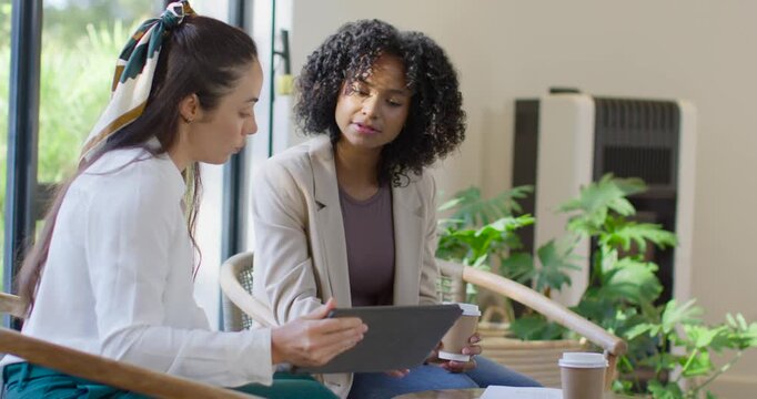 Diverse women colleagues in office lounge, left woman sharing tablet, tapping, reviewing documents