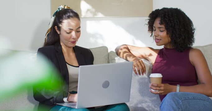 Two adult women sitting on sofa, left in blazer placing laptop while right holding cup, reviewing