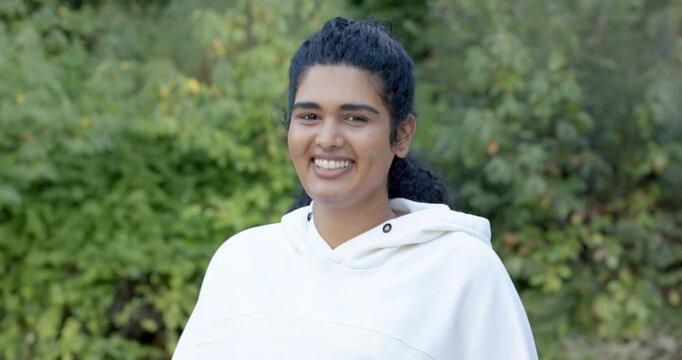 Woman 20s responding to camera prompt, blinking, widening smile, leaning in garden with hoodie