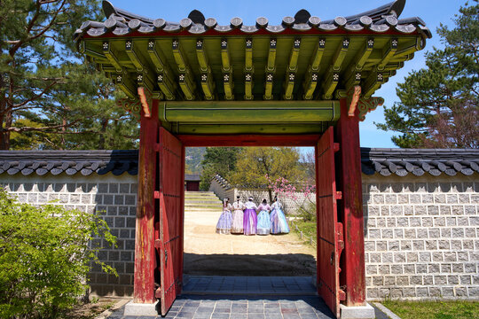 Traditional wooden gate of Gyeongbokgung Palace in Seoul, South Korea, featuring gracefully curved hanok-style roof, wide open to reveal 5 young women in vibrant hanbok. Dressed in flowing silk skirts