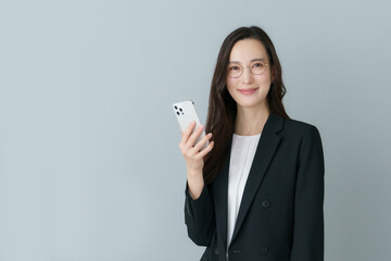 Fototapeta premium グレーバックの眼鏡をかけたスマホを持つ若いスーツの日本人女性