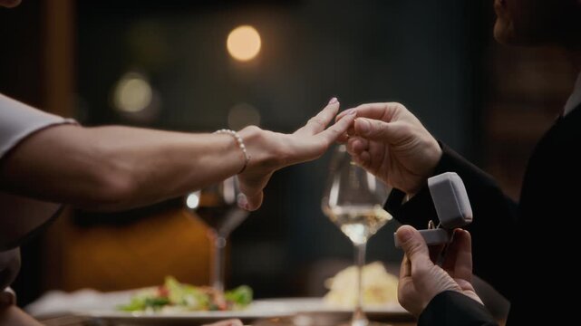 Cropped shot of unrecognizable man in suit presenting engagement ring and proposing to happy elegant woman during restaurant dinner with wine glasses in blurred background