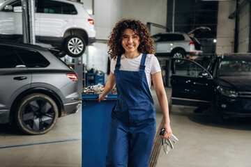 Female auto mechanic with curly hair wearing blue overalls stands beside a workbench holding tools in a busy automotive garage with cars in the background © standret