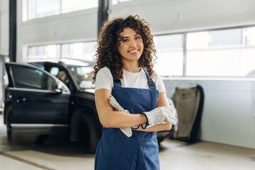 Female auto mechanic with curly hair wearing blue overalls and gloves stands confidently in a garage with a car in the background and bright natural light © standret