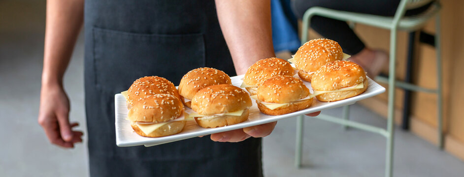Banner of unrecognizable waiter in a black apron holding a white tray with delicious mini burgers with sesame seed buns and cheese, offering appetizer snacks at a catered event or restaurant