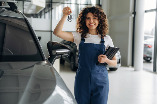 Female auto sales representative in blue overalls holds car keys and tablet, standing beside a gray vehicle in a modern car dealership showroom