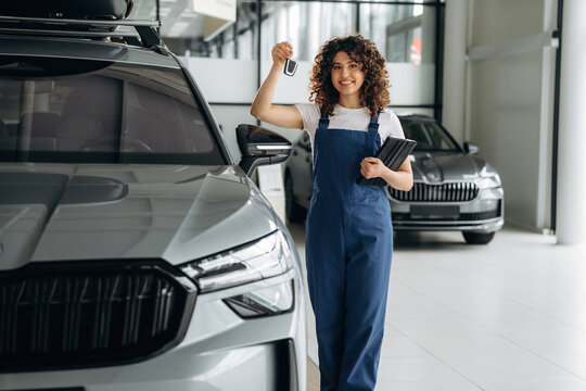 Female auto sales representative in blue overalls holds car keys and tablet, standing beside a silver vehicle in a modern car dealership showroom with additional cars visible
