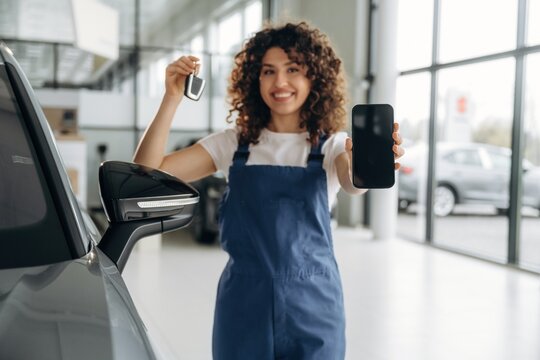 Female auto mechanic in blue overalls holds car keys and smartphone, standing next to a vehicle inside a modern automotive showroom with large windows