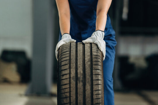 Female auto mechanic in blue coveralls lifts a car tire with both hands inside a garage, showcasing tools and equipment in the background